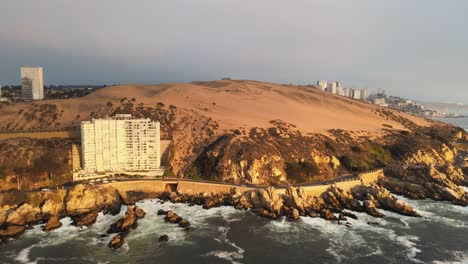 Aerial-dolly-out-of-orange-sand-dunes-in-rugged-hillside-next-to-sea-waves,-Concon-apartments-and-resorts-on-an-overcast-day,-Chile
