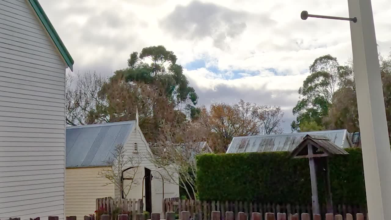 A quaint wooden structure with green accents, surrounded by trees and a wooden fence under a cloudy sky.