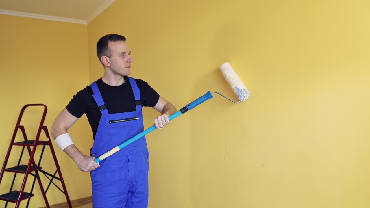 Room renovation at home. Builder specialist painting yellow wall into new color. Portrait of a young craftsman in overalls starts painting wall with a paint roller.