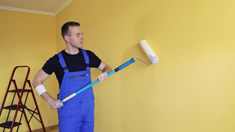 room renovation at home. builder specialist painting yellow wall into new color. portrait of a young craftsman in overalls starts painting wall with a paint roller.