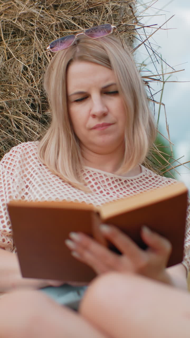 Primo piano di una donna con un top a rete e occhiali da sole appoggiati sulla testa, assorta nella lettura di un libro, si appoggia a una balla di fieno in un campo aperto con alberi e cielo sfocati.