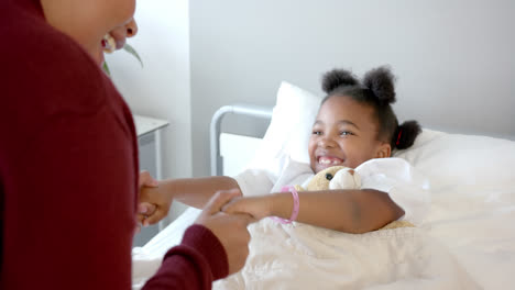 happy african american mother fooling with daughter lying in hospital bed with mascot, slow motion