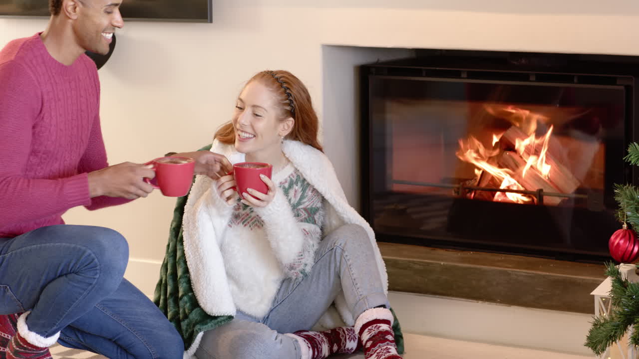 Diverse couple enjoying hot cocoa by cozy fireplace during Christmas