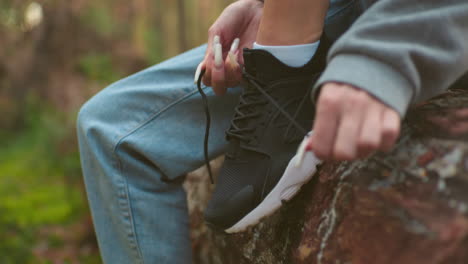 close-up of woman with long white nails tying shoelaces on black sneakers, wearing gray sweater and light blue jeans, seated on fallen tree during hike in forest