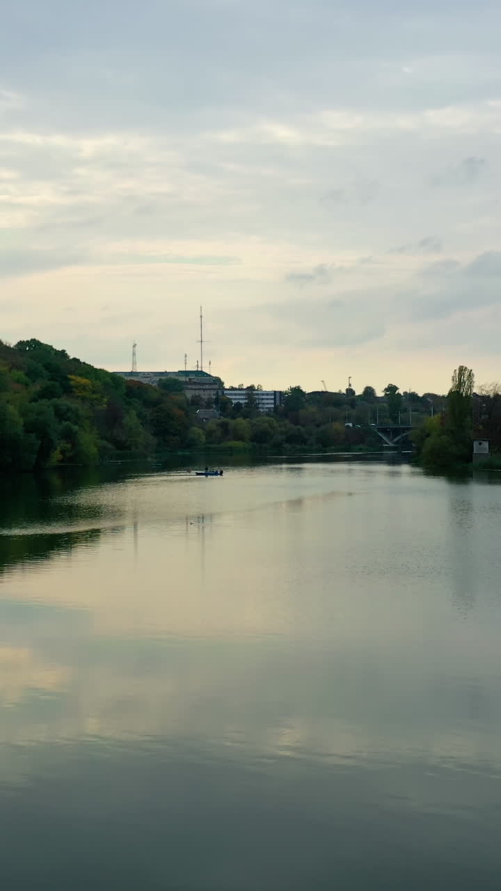 Calm water in the evening. River among green trees from both sides. Panoramic view of a river in the town. Motion camera back. Aerial view. Vertical video