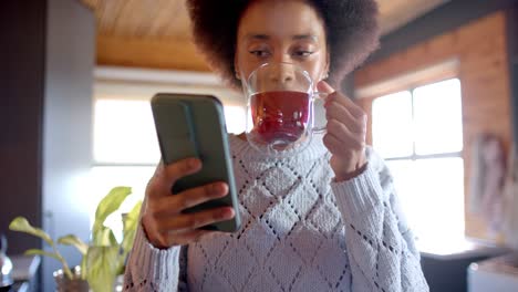 happy african american woman using smartphone and drinking tea in sunny kitchen, in slow motion