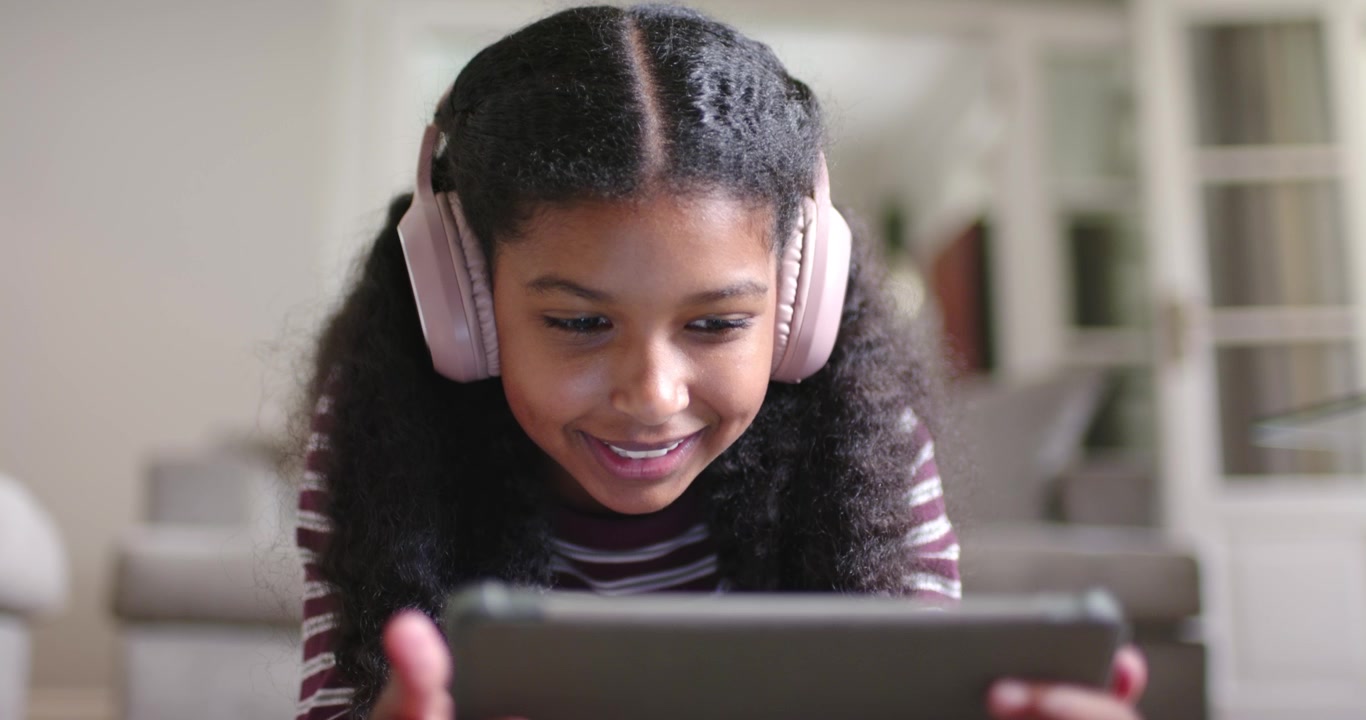 Afroamerikansk teenager reagerer overrasket på en tablet, flytter sig, smiler med lyserødt headset på sofaen