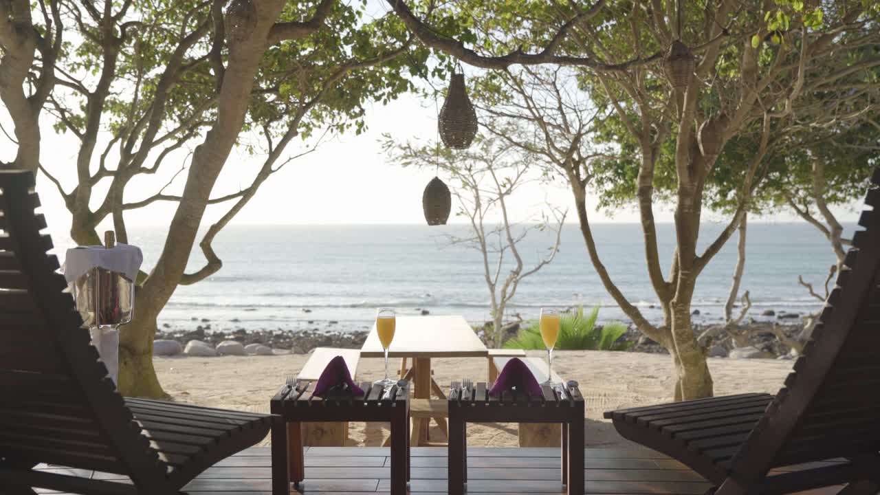 Static scene from a luxury resort seeing the ocean between two lounge chairs with glasses of mimosas and bottle of champagne on the side