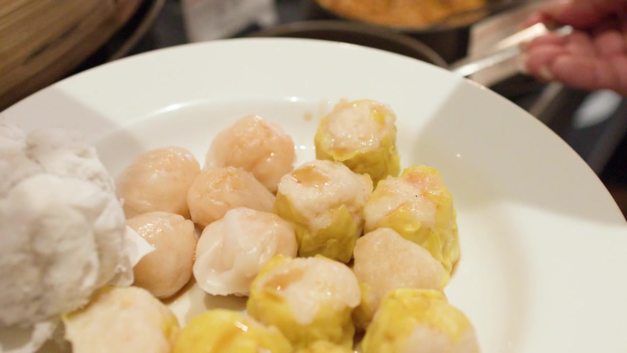 Sendok logam menuangkan kecap hitam di atas berbagai shumai dan pangsit kukus di piring putih.