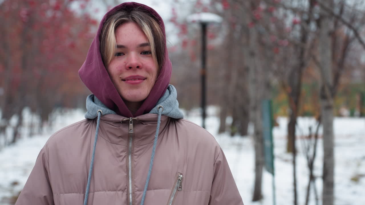 따뜻한 후드 재을 입은 젊은 여성은 눈 인 길, 은 베리 나무와 먼 거리 램프와 함께 겨울 공원 설정에 서있는 동안 주머니에 손으로 미소