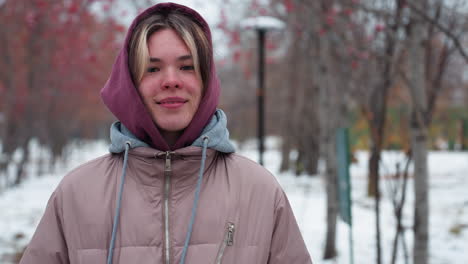 mujer joven con una chaqueta con capucha cálida sonríe con las manos en los bolsillos mientras está de pie en el camino cubierto de nieve, el parque de invierno con árboles de bayas rojas y farolas lejanas