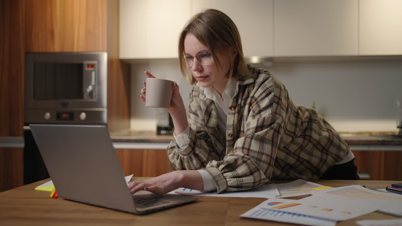 A young beautiful business woman works from home with a cup of coffee and looks into a laptop screen standing at the table. View mail and drink coffee. Remote work from home