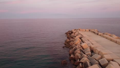 low aerial forward of long rocky jetty in blue ocean sea during purple sunset