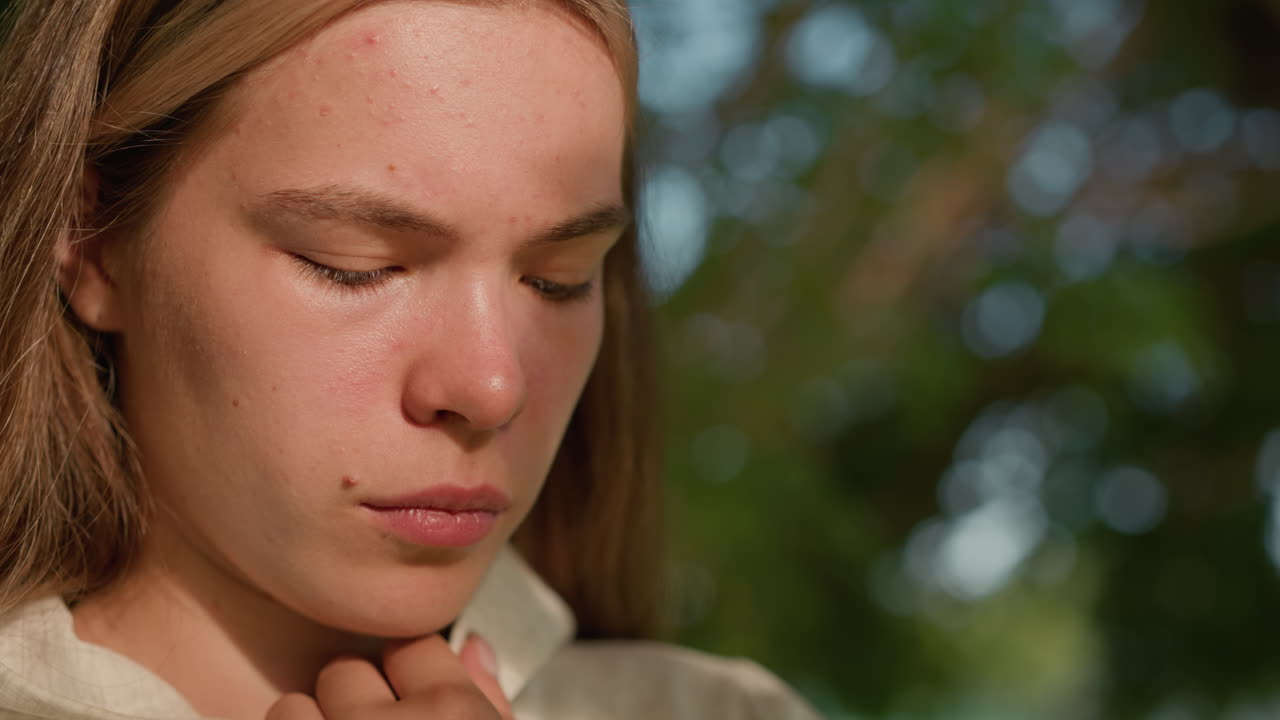 Close-up of young woman in deep thought holding hood collar, face softly illuminated by sunlight filtering through leaves, with background greenery blurred