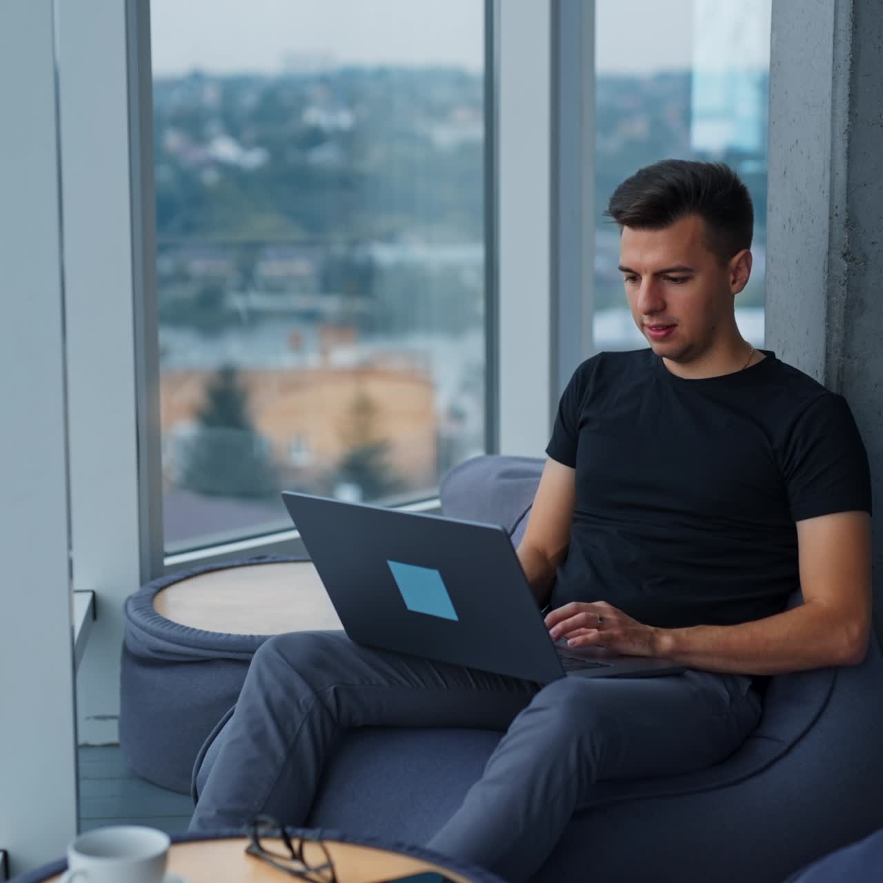Dark-haired Caucasian man sitting in bean bag chair works on laptop. Male office employee at work. City at backdrop in blur