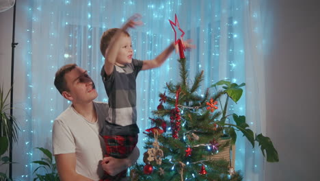 Dad-lifted-the-boy-to-the-top-of-the-Christmas-tree-to-place-a-Christmas-star-on-top-of-the-top.-Father-and-son-decorate-Christmas-tree-together.-High-quality-4k-footage