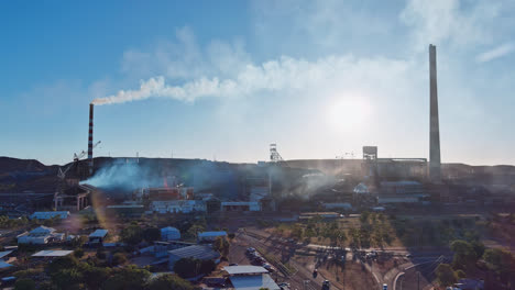 Track-back,-Mount-Isa-mines-with-smoke-plumes-from-its-chimneys-with-cars-driving-and-buildings-in-the-foreground,-Mount-Isa,-Queensland,-Australia