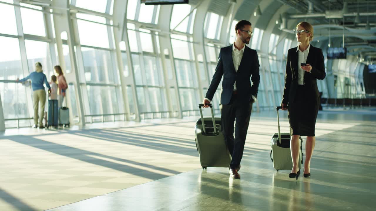 Business Couple Of The Co Workers Travelling On The Working Trip, Walking The Airport With Suitcases On Wheels And Talking