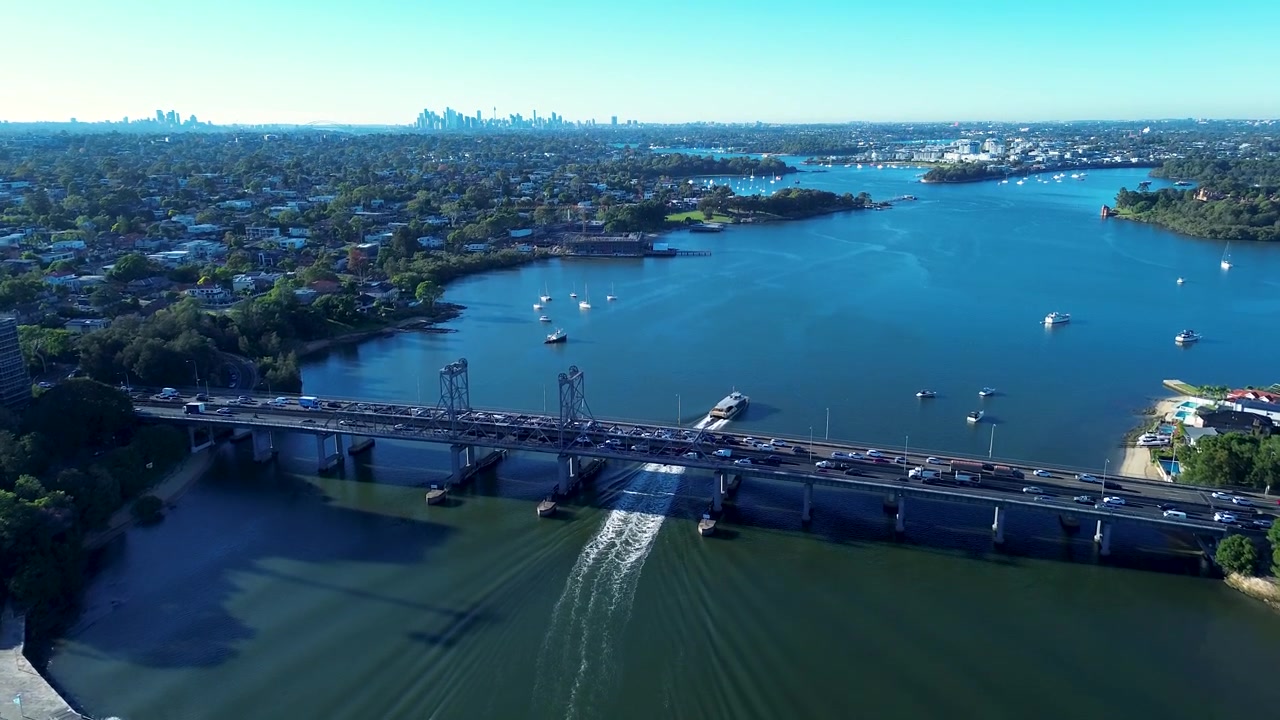 Paesaggio aereo con drone di un traghetto pendolare che naviga lungo il fiume Parramatta con traffico automobilistico che attraversa il ponte Ryde e lo skyline della città di Sydney sullo sfondo vicino a Meadowbank, NSW, Australia. Trasporto.