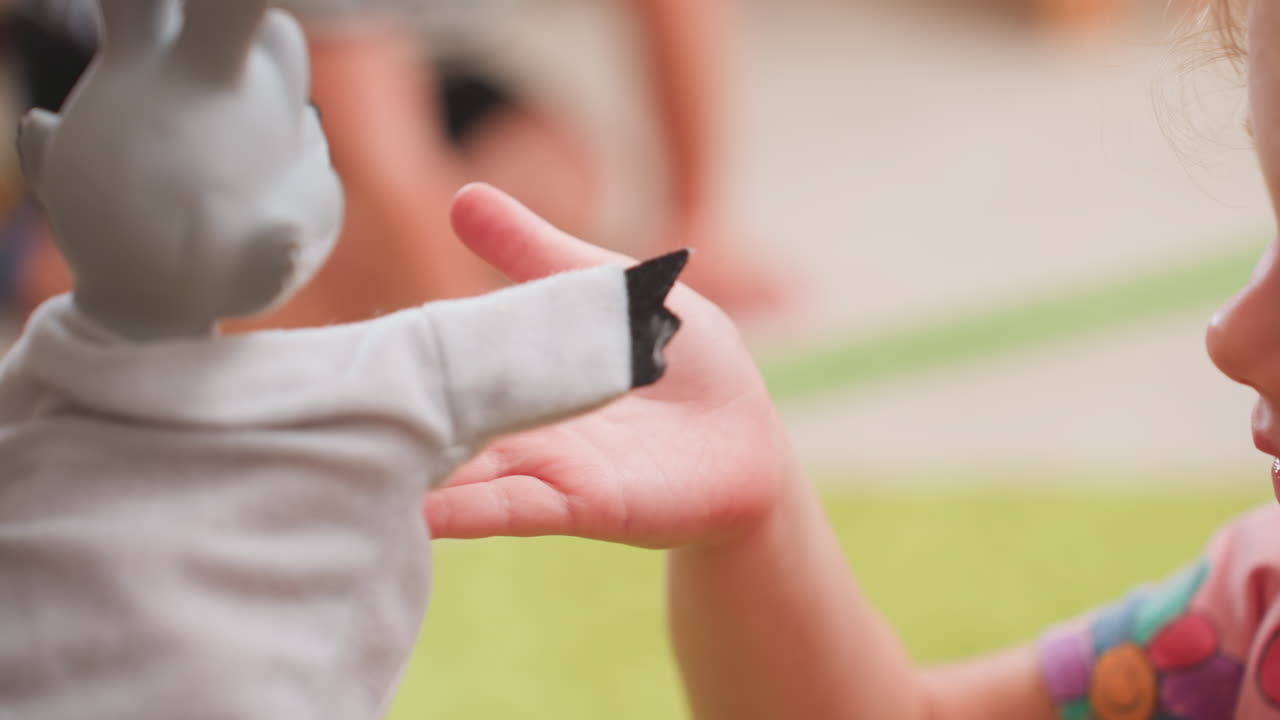 Close up shows toddler hand extended for puppet touch during classroom play, encouraging sensory interaction, trust, and communication while blurred child in stripe clothing moves away in background