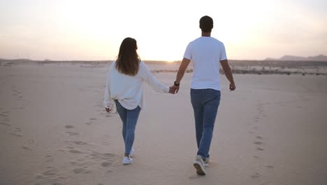 Backside-footage-of-holding-hands-couple-walking-in-romantic-relationship-under-sun-and-blue-sky-in-desert.-Two-young-lovers-walking-by-desert-sand-in-casual-clothes.-Landscape-on-background.-Slow-motion