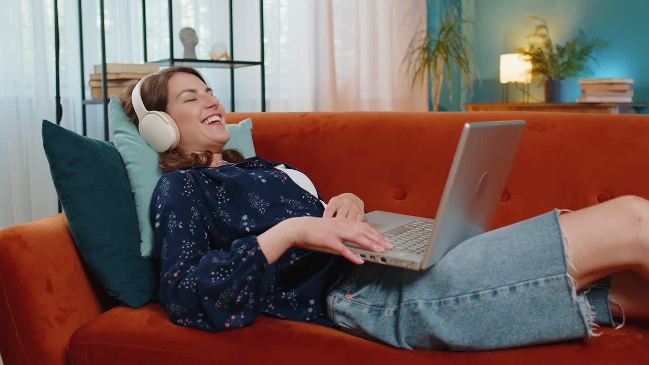 Happy woman in wireless headphones lying on home couch listening music working on laptop relaxing