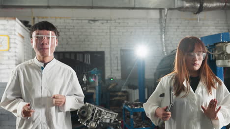 dos estudiantes con abrigos de laboratorio blancos y gafas protectoras bailan felices juntos en un taller mecánico, mostrando un momento enérgico y lúdico en medio del entorno industrial