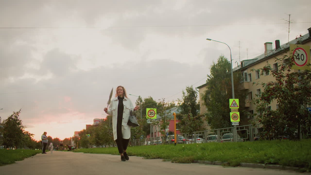 woman walking along paved walkway carrying handbag and twirling umbrella joyfully with cars passing in distance and people standing near residential buildings and parked vehicles during sunset