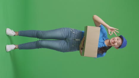 full body of asian female courier in blue uniform showing okay gesture and smiling while delivering a carton on green screen background in the studio