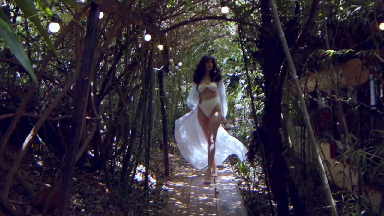 Splendida modella in bikini bianco e vestito bianco cammina attraverso una giungla al rallentatore.
