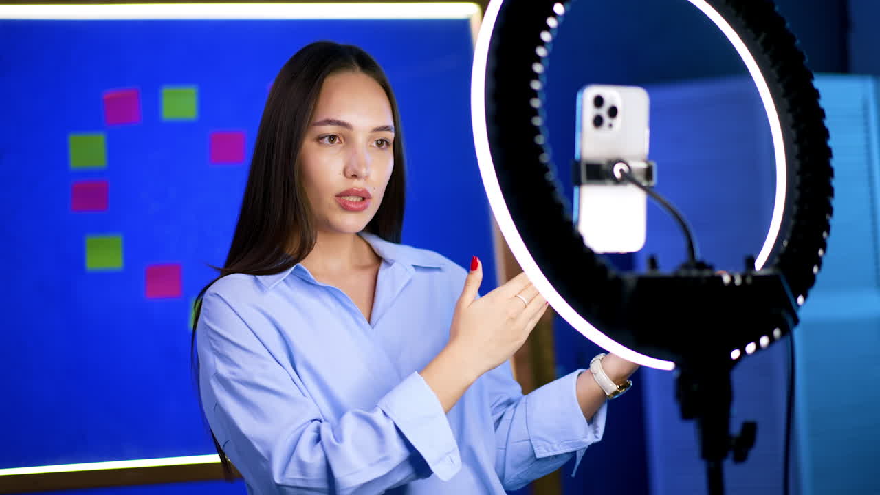 Girl in a blue shirt stands in front of the light ring and phone on tripod. Lady talking for her blog video.