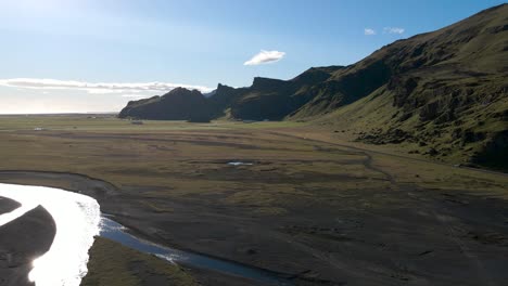 A-stunning-contrast-of-Iceland's-landscape,-where-flat-terrain-meets-a-towering-green-mountain-beside-the-road,-all-under-the-brilliant-sunshine,-captured-in-4K