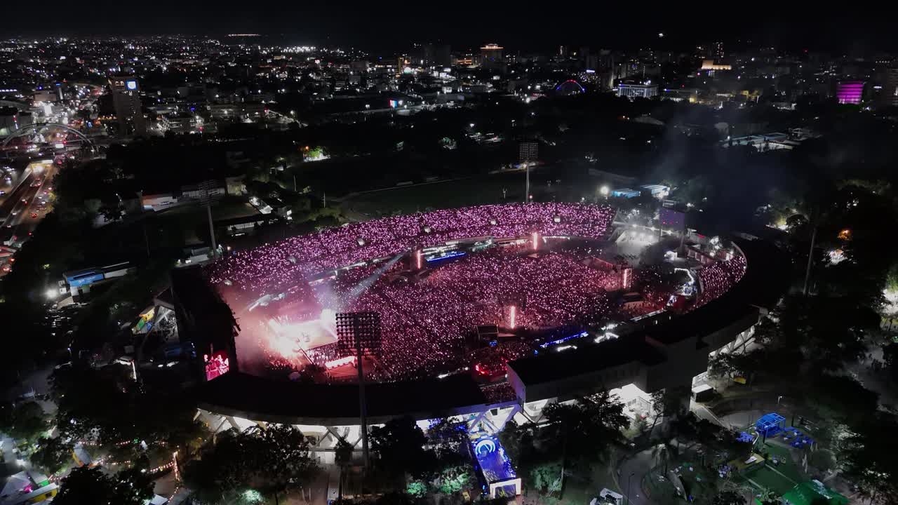 Aerial approaching shot showing crowd of people celebrating bad bunny festival show in open air stadium at night. Santo Domingo, Dominican Republic. Music show at night with flashing illuminated light