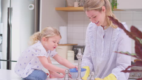 mother wearing rubber gloves at home in kitchen with young daughter having fun as they do washing up at sink- shot in slow motion