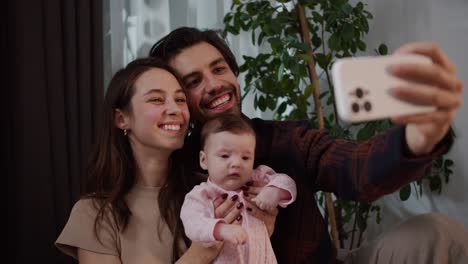 happy young brunette guy with stubble together with his brunette wife play with their little daughter baby in a pink suit and take a family selfie photo using a white smartphone in a modern apartment