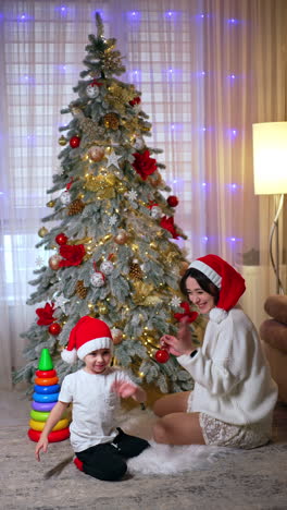 happy smiling woman in red santa cap sits near the christmas tree. little kid crawls by the floor nearby. vertical video.