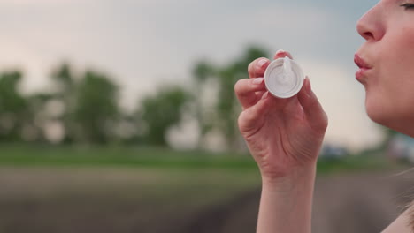close up of woman lips and hand holding bubble wand dripping soap solution as gentle breeze creates iridescent bubbles against soft focus farmland backdrop at golden hour