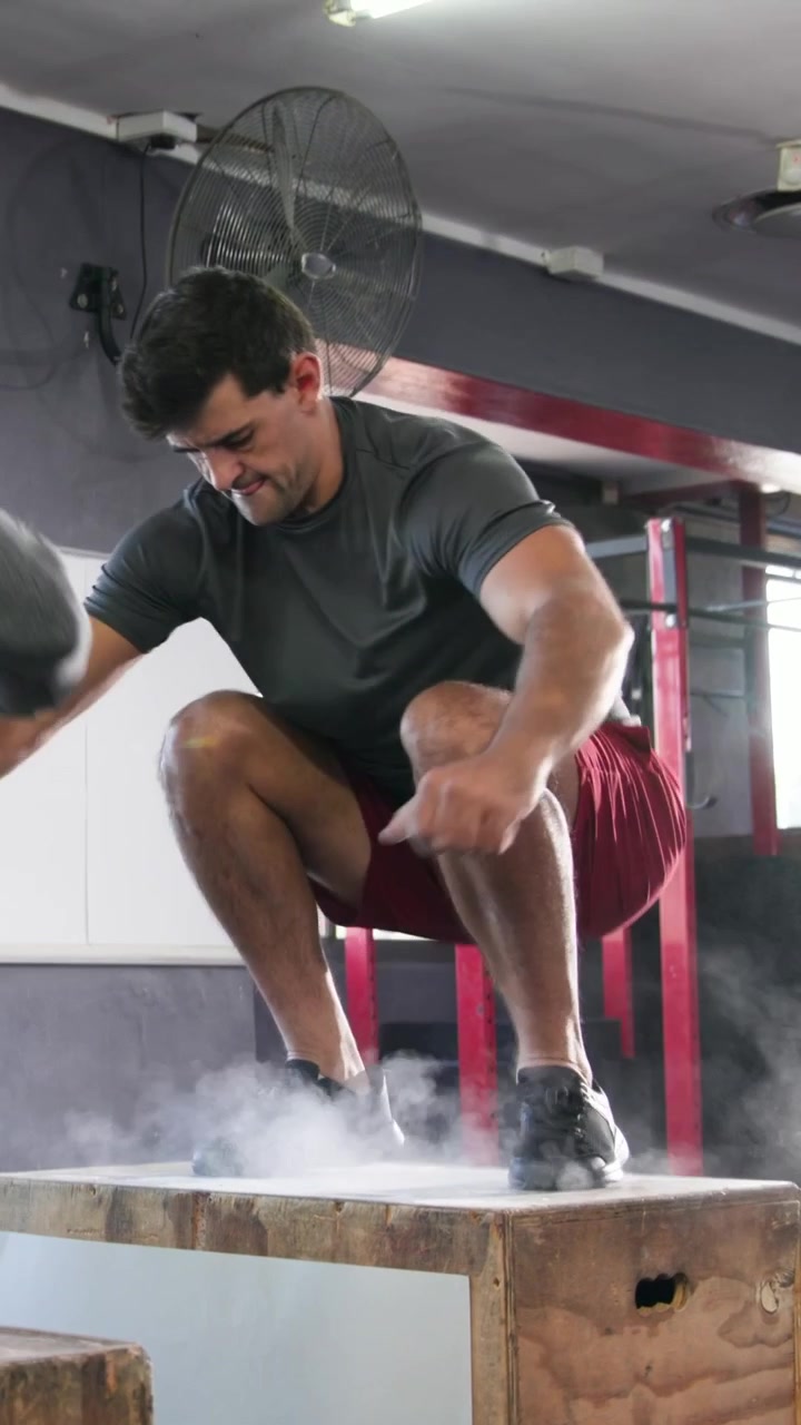 Vídeo vertical: Atleta de shorts vermelhos se aproximando da caixa pliométrica, fazendo saltos em caixa para potência na academia, giz.
