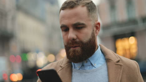 Vista-Cercana-Del-Hombre-De-Negocios-Caucásico-Con-Barba-Enviando-Mensajes-De-Texto-Por-Teléfono-En-La-Calle-En-Otoño