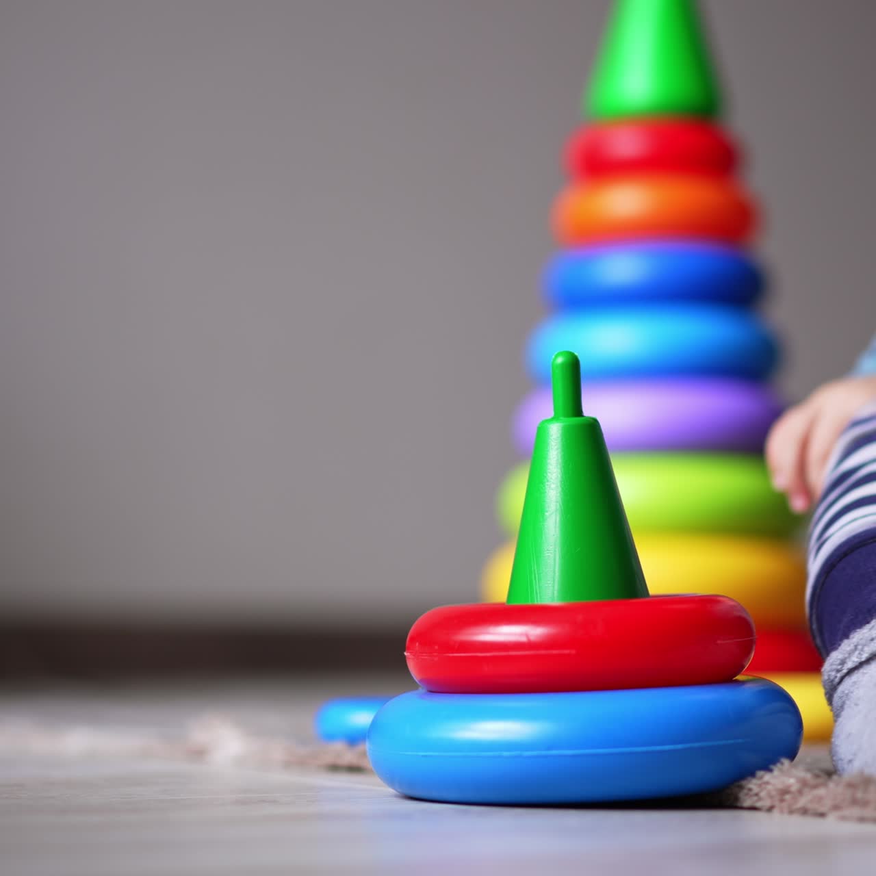 Small baby playing with little toy pyramid on the floor. Kid put the ring on and off the basis. Bigger pyramid at backdrop in blur
