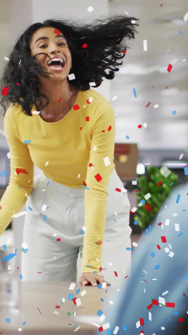 Video verticale: Coriandoli che esplodono, donna che si china su un tavolo da lavoro, raggiungendo un foglio, festeggiando.
