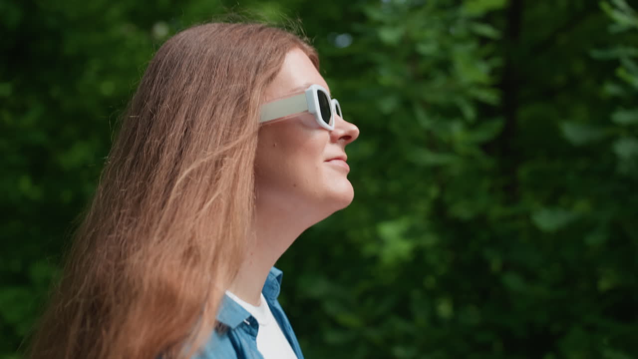 Primer plano de una mujer paseando en un día soleado con gafas de sol blancas y camisa azul, sonriendo suavemente mientras disfruta de la cálida luz del sol y un fondo de naturaleza verde