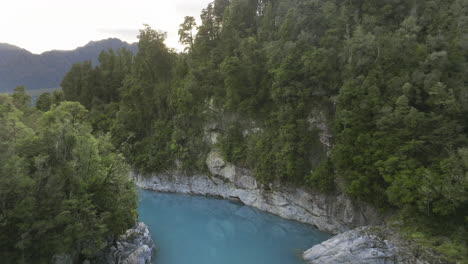 Volando-Más-Allá-Del-Puente-Colgante-Que-Cruza-El-Famoso-Río-Azul-Del-Desfiladero-De-Hokitika,-Aéreo