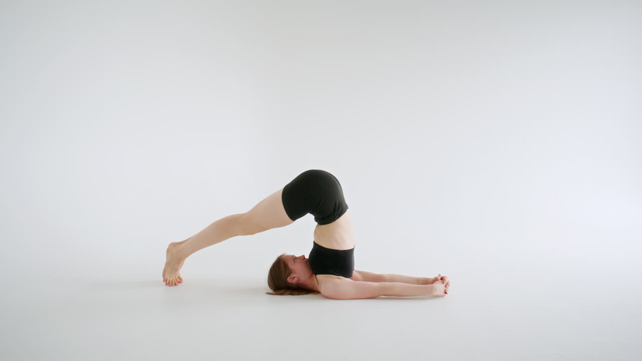Frau übt Halasana-Pose auf dem Boden in einem weißen Yoga-Studio