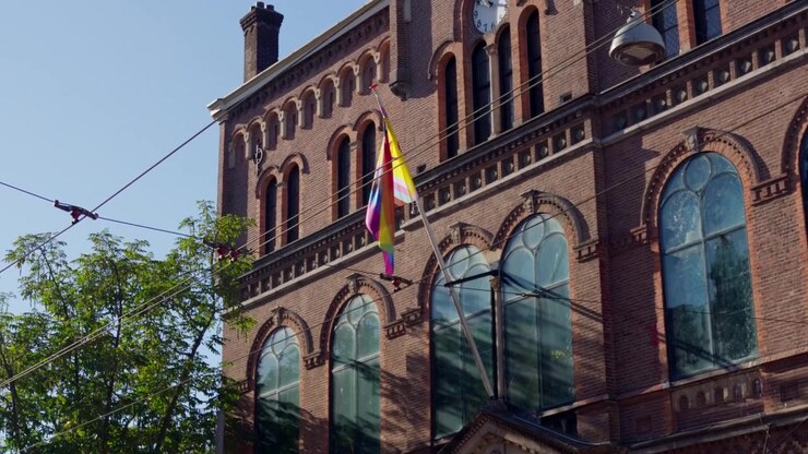 Dettaglio dal basso di un esterno in mattoni storici, finestre ad arco e una bandiera inclusiva del pride che sventola montata sul famoso locale di musica Paradiso sotto un cielo azzurro limpido del mattino