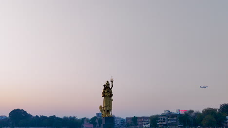 Behold-the-splendour-of-a-beautiful-front-view-of-the-immense-gilded-Lord-Shiva-statue-at-Sursagar-Lake-in-Vadodara-at-dusk,-with-a-low-flying-plane-passing-in-the-background