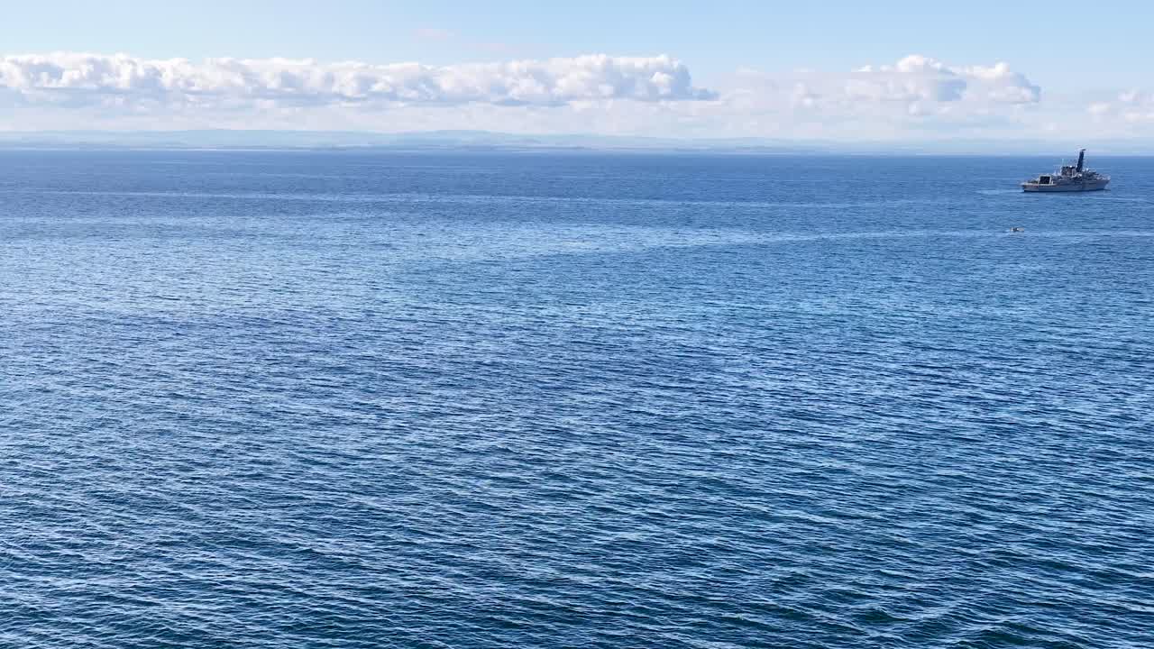 Wide shot of naval ship moving past rugged coastal cliffs, bright daylight, gentle camera pan