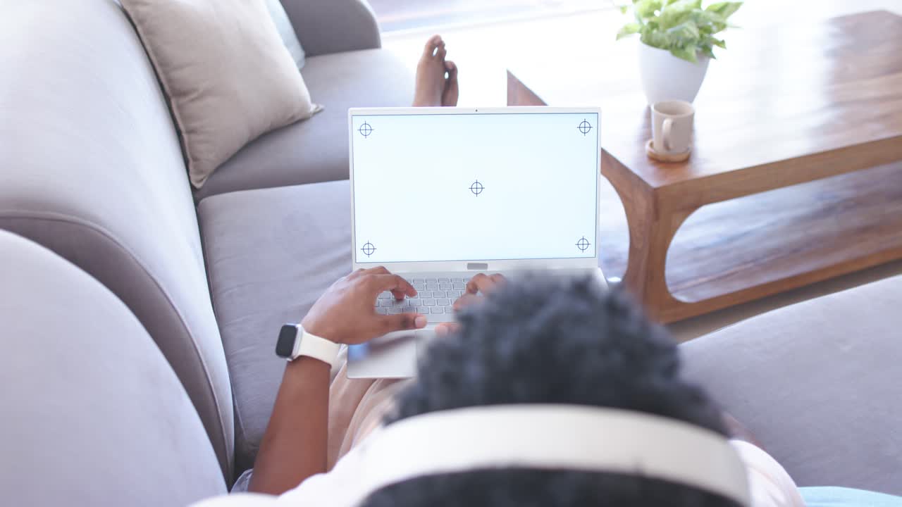 African American man reclining on couch with headset, shifting forward, typing on laptop, working