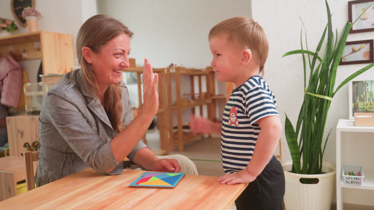 Una donna sorridente dà il cinque a un bambino allegro con apparecchio acustico dopo aver completato un'attività con un puzzle colorato su un tavolo di legno, celebrando il successo con gioia condivisa in un ambiente di apprendimento luminoso