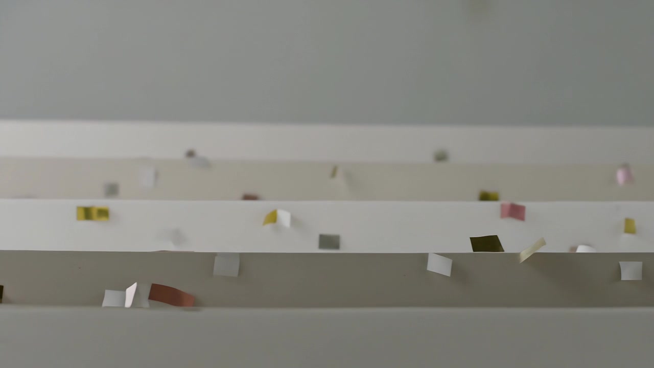 Drifting confetti pieces driven by gravity and breeze, catching on white slats at pale gray wall
